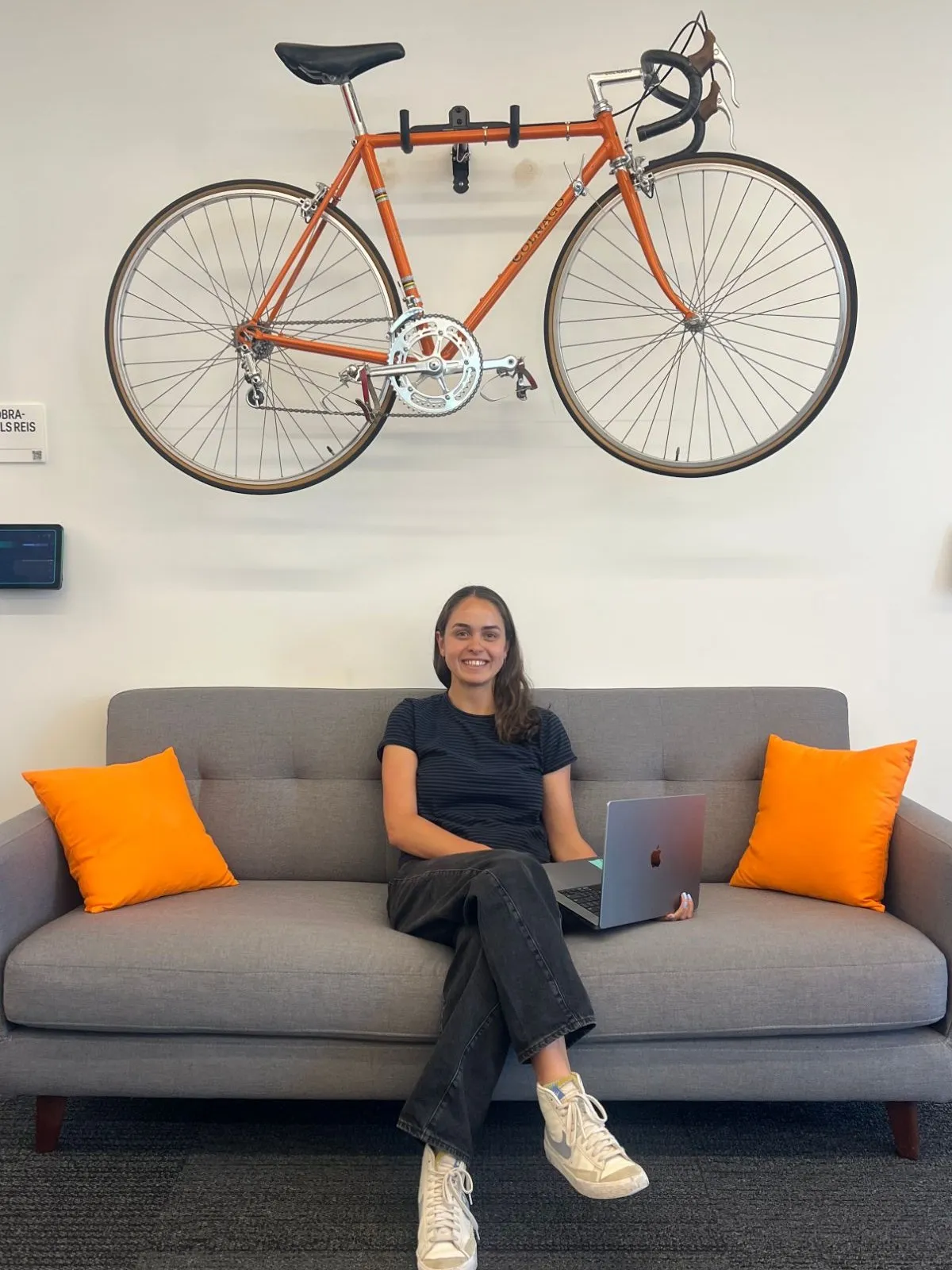 Grace Melcher smiling on a gray couch with a laptop, under an orange road bike mounted on the wall
